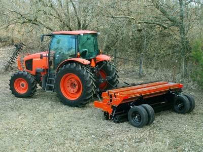 A side view of an orange Land Pride NTS2509 Seeder driving on grass with trees behind it